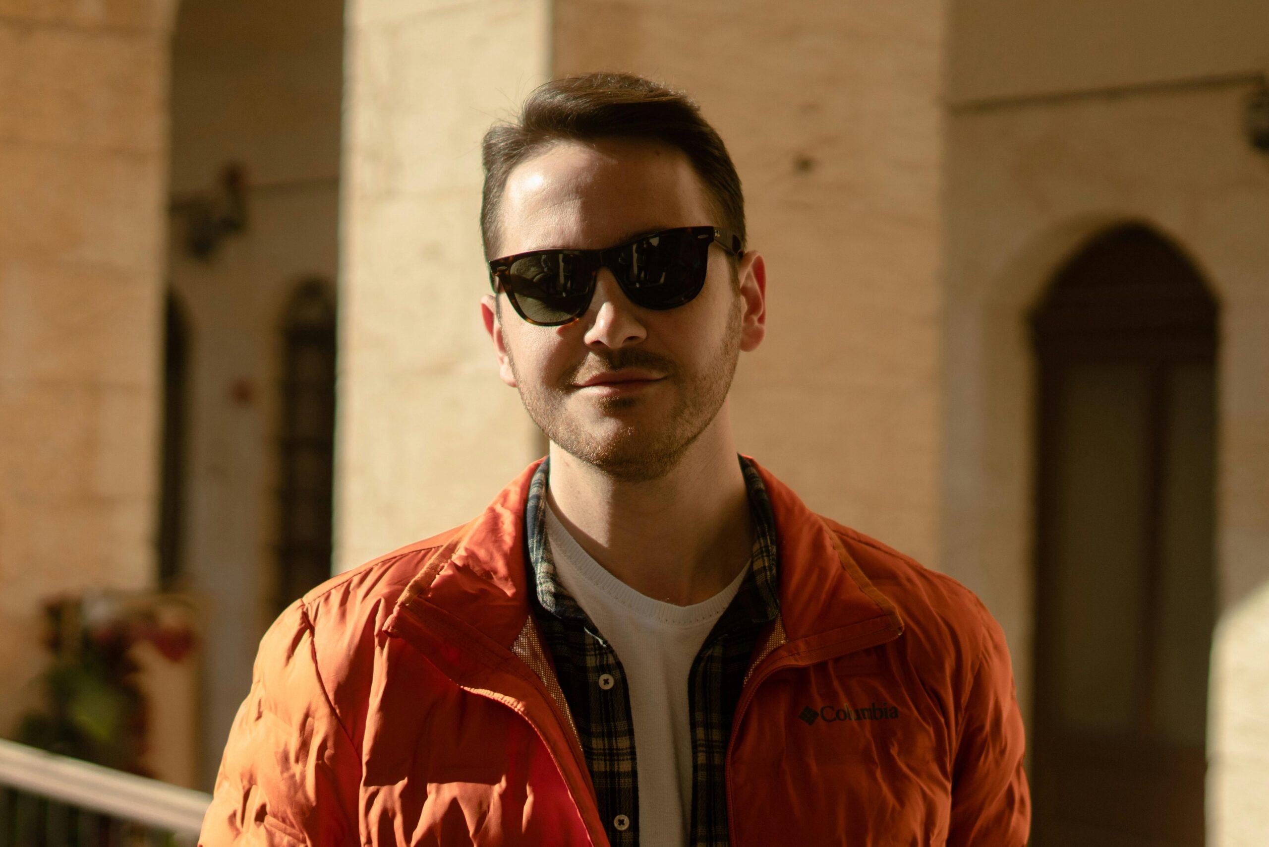 Close-up portrait of an adult man wearing sunglasses and an orange jacket indoors.
